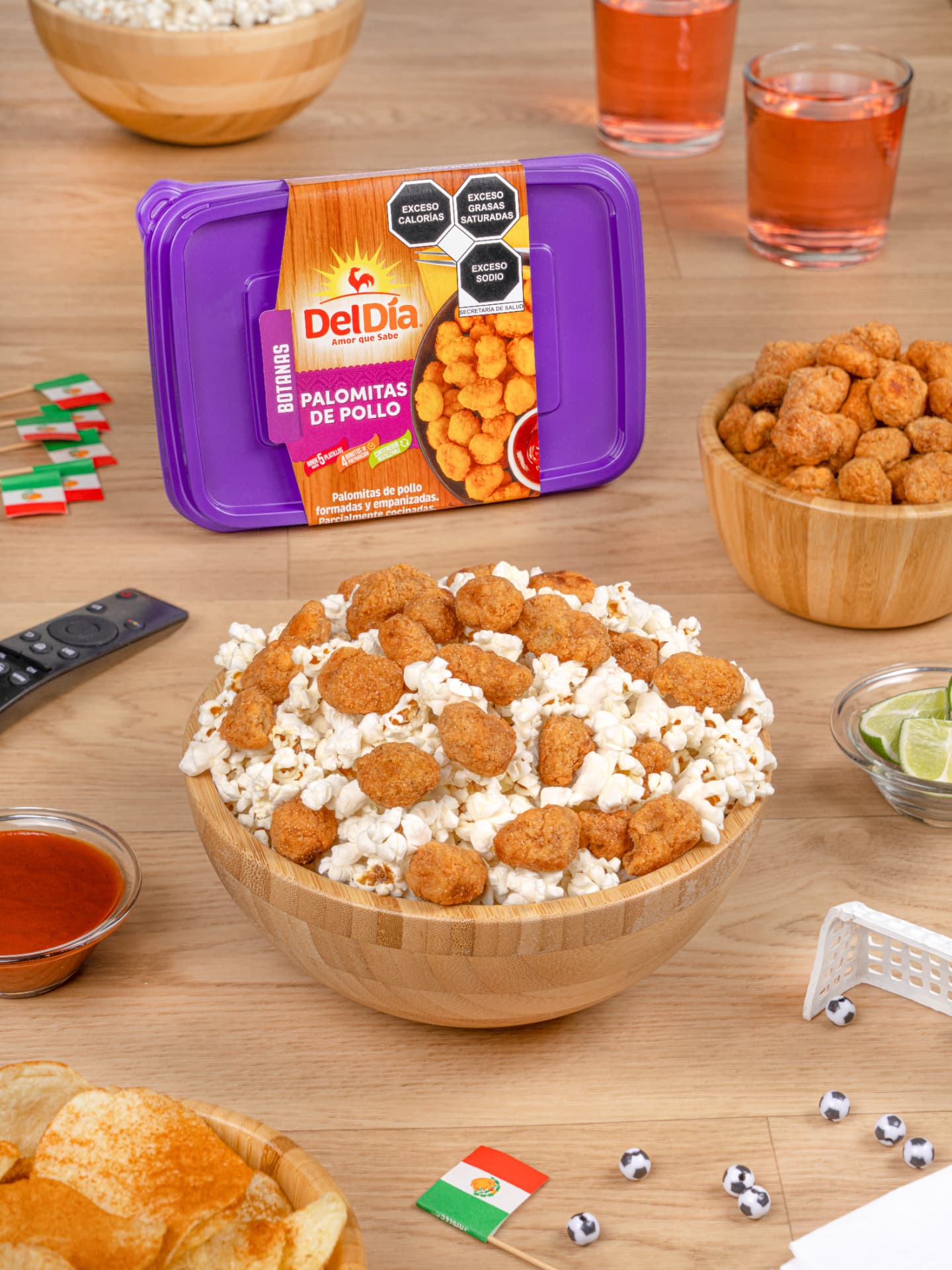 Palomitas de pollo DelDía en bowl de bambú con botanera, papas, salsa roja, limones y detalle de fútbol sobre mesa de madera, fotografía vertical
