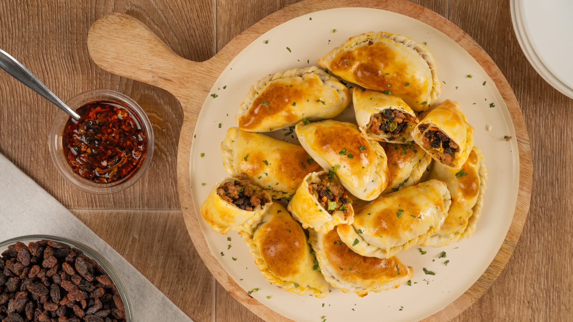 Empanadas de carne con pasitas cortadas a la mitad sobre un plato crema, con salsa de chile y un tazón de pasitas, vista cenital