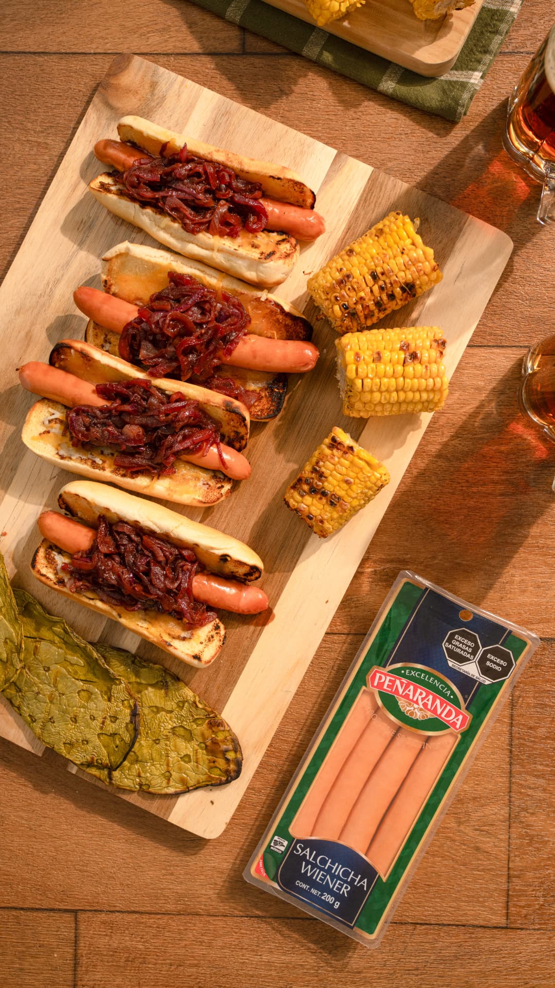 Hot dogs con cebolla caramelizada, elote y nopales asados sobre tabla de madera, junto a un paquete de salchicha Wiener Peñaranda y dos cervezas, vista cenital