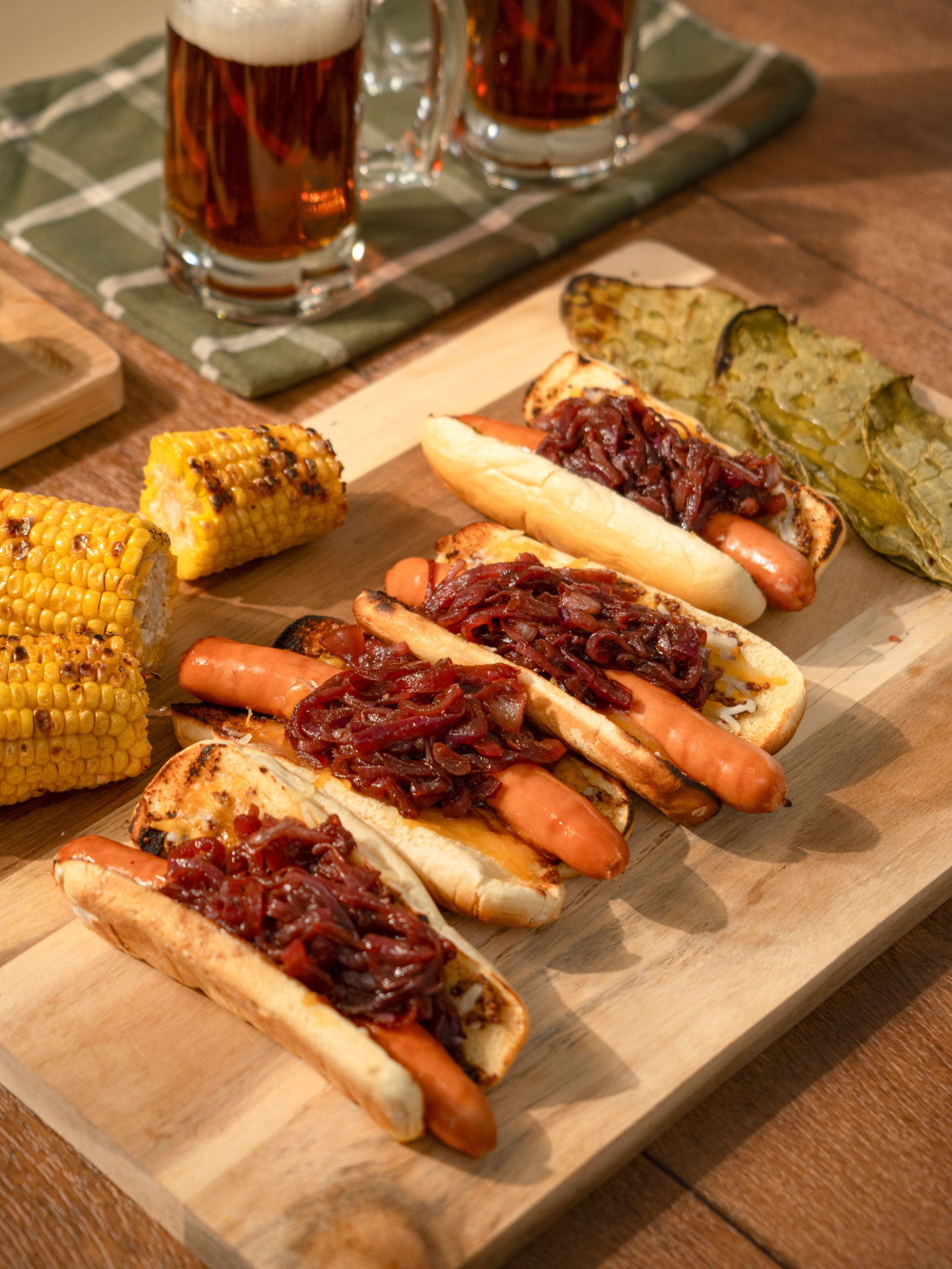 Hot dogs con cebolla caramelizada, elote y nopales asados sobre tabla de madera, vista diagonal