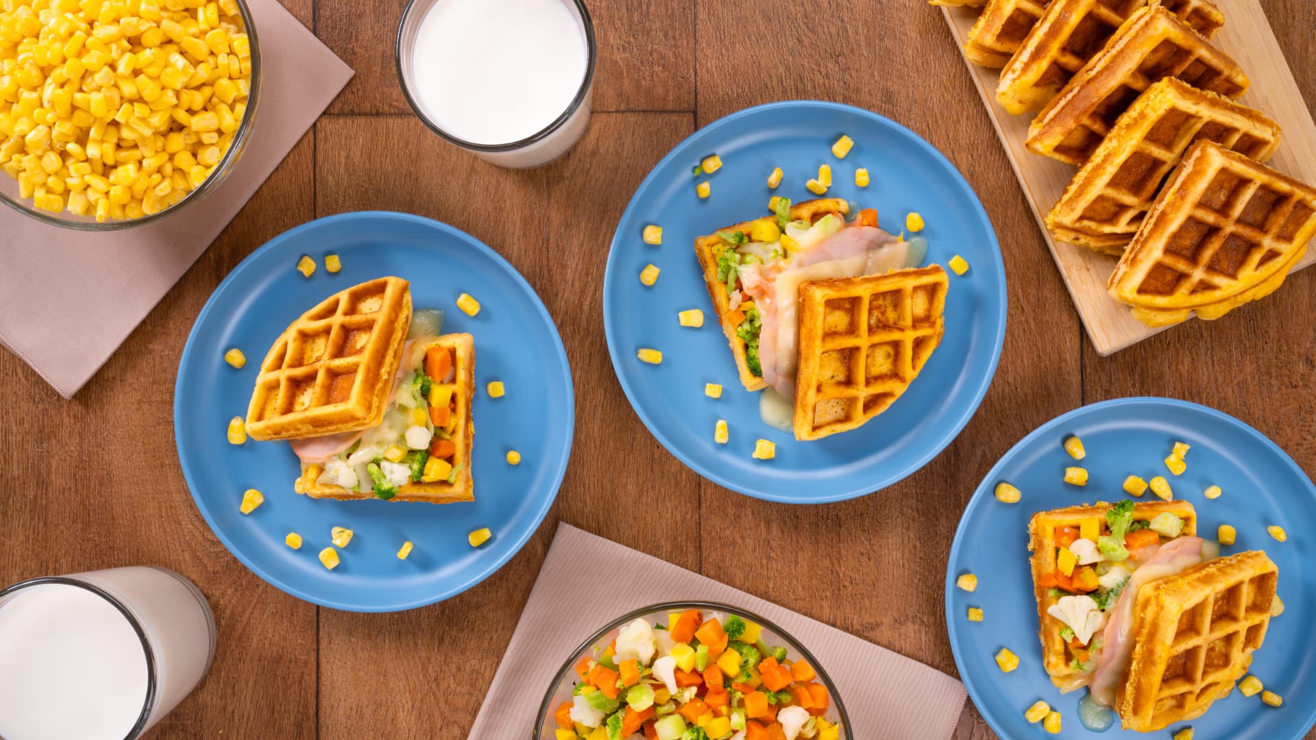 Sándwiches de waffles con jamón, verduras y elote en platos azules, con waffles, leche y maíz alrededor, vista cenital