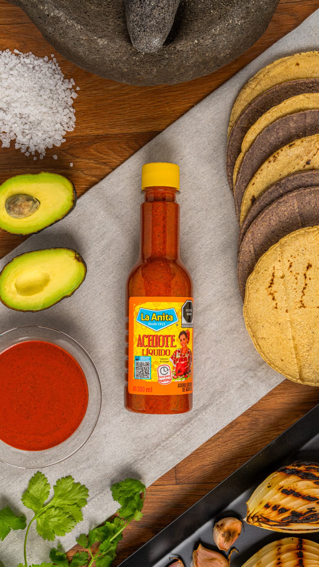 Botella de achiote líquido La Anita acompañada de molcajete, aguacates, tortillas y cilantro sobre mesa de madera