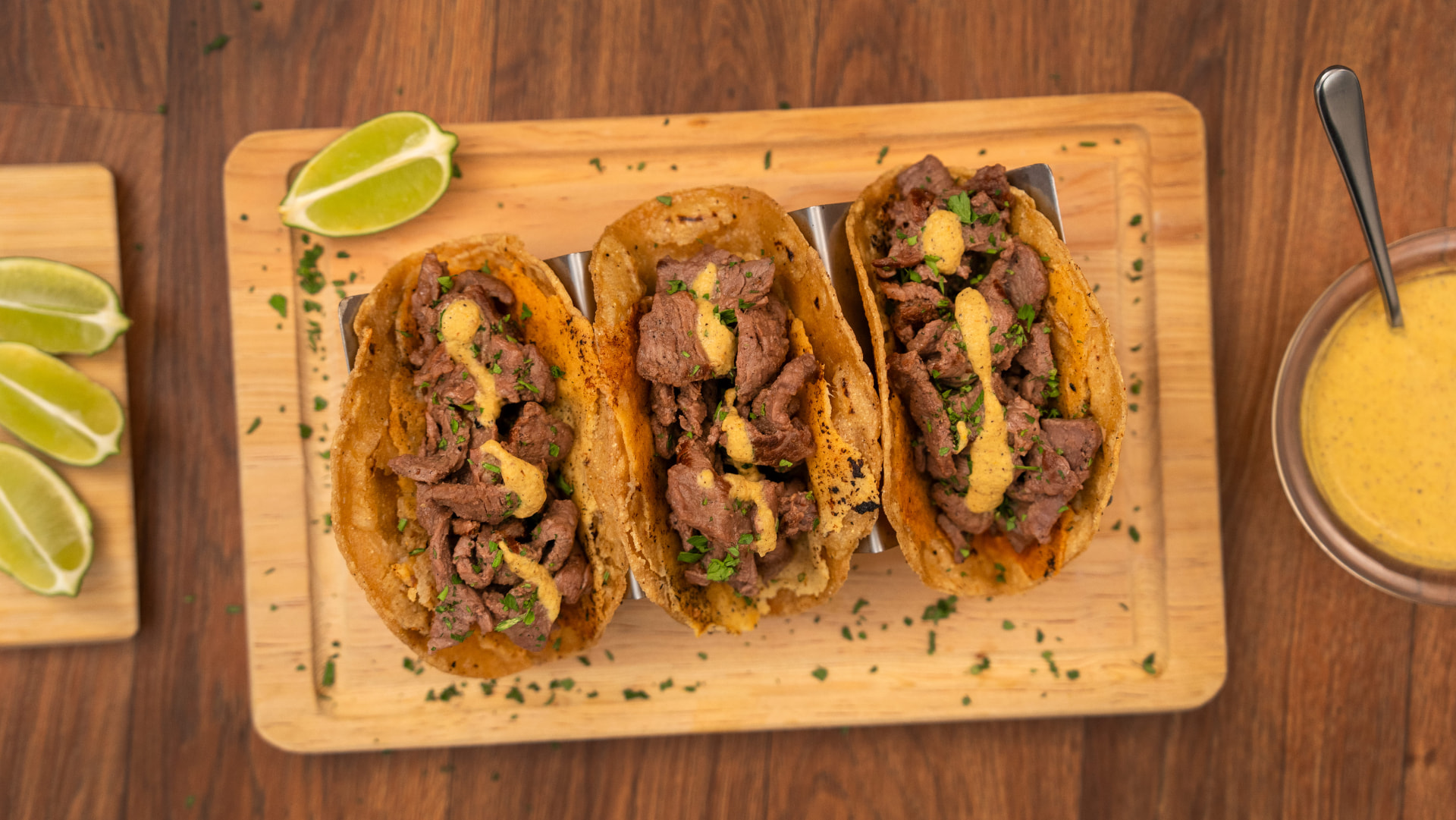 Tacos de sirloin con crema amarilla y hierbas picadas en soporte de metal sobre tabla de madera, vista cenital